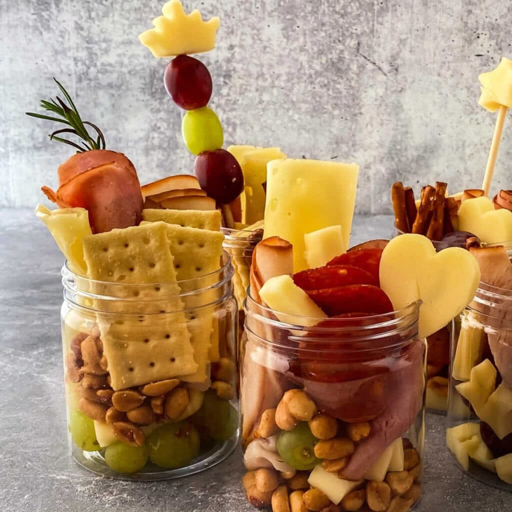 Charceuterie in jars on a table with fruit skewers.