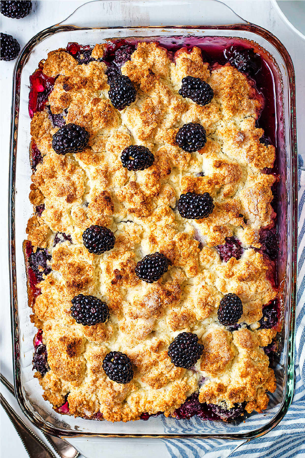 A freshly baked blackberry cobblre in a glass dish with blackberries scattered on top.
