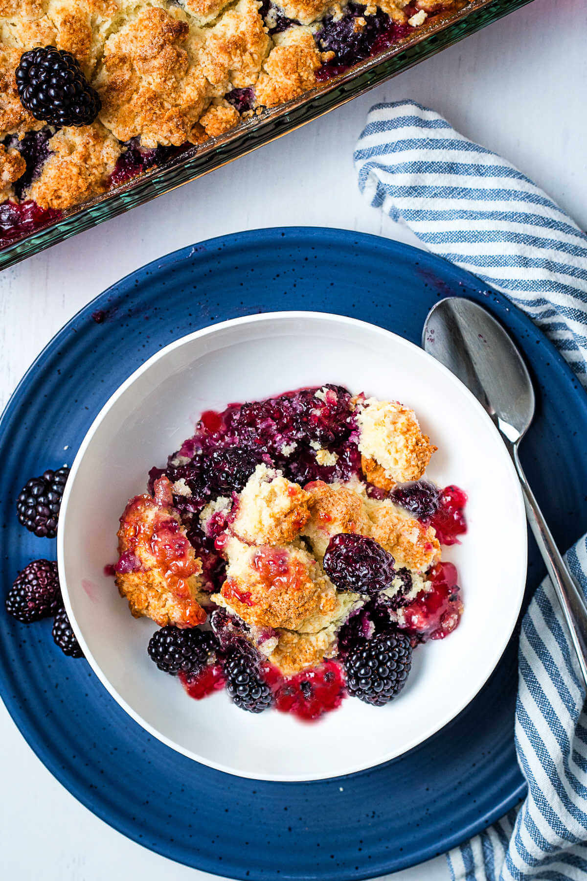 A serving of blackberry cobbler in a bowl on a table with a baking dish of cobbler in the background.
