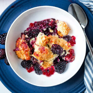 A bowl of blackberry cobbler on a plate with a spoon and napkin.