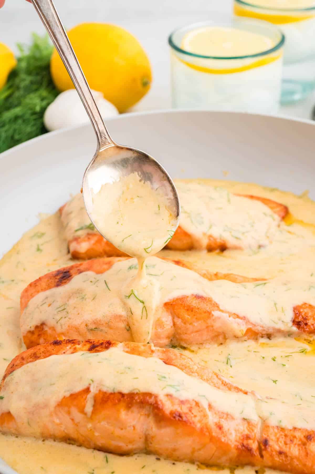Spooning dill cream sauce over salmon filets in a pan.