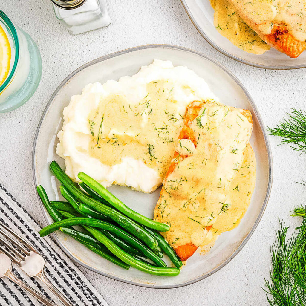 A dinner plate on a table with salmon with dill cream sauce, mashed potatoes, and green beans.