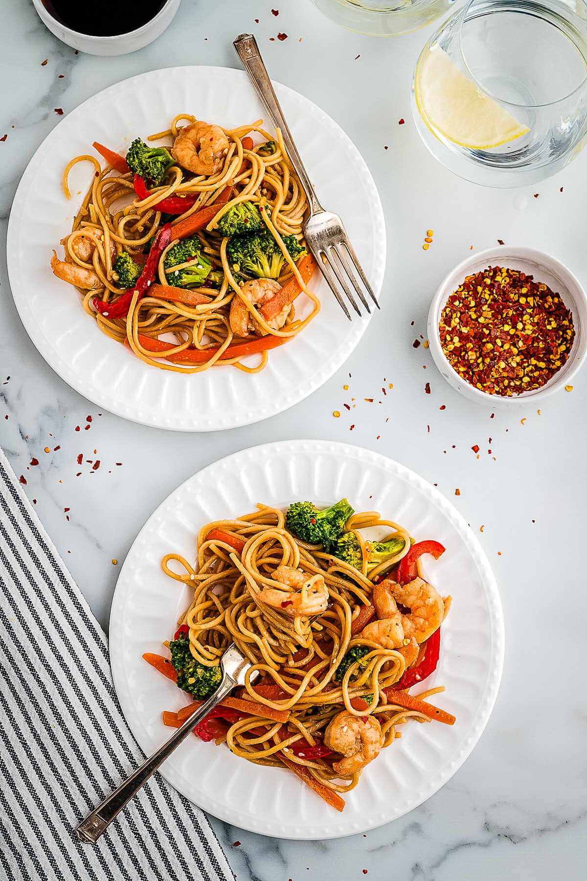 Two servings of shrimp lo mein on plates on a table with a bowl of red pepper flakes.