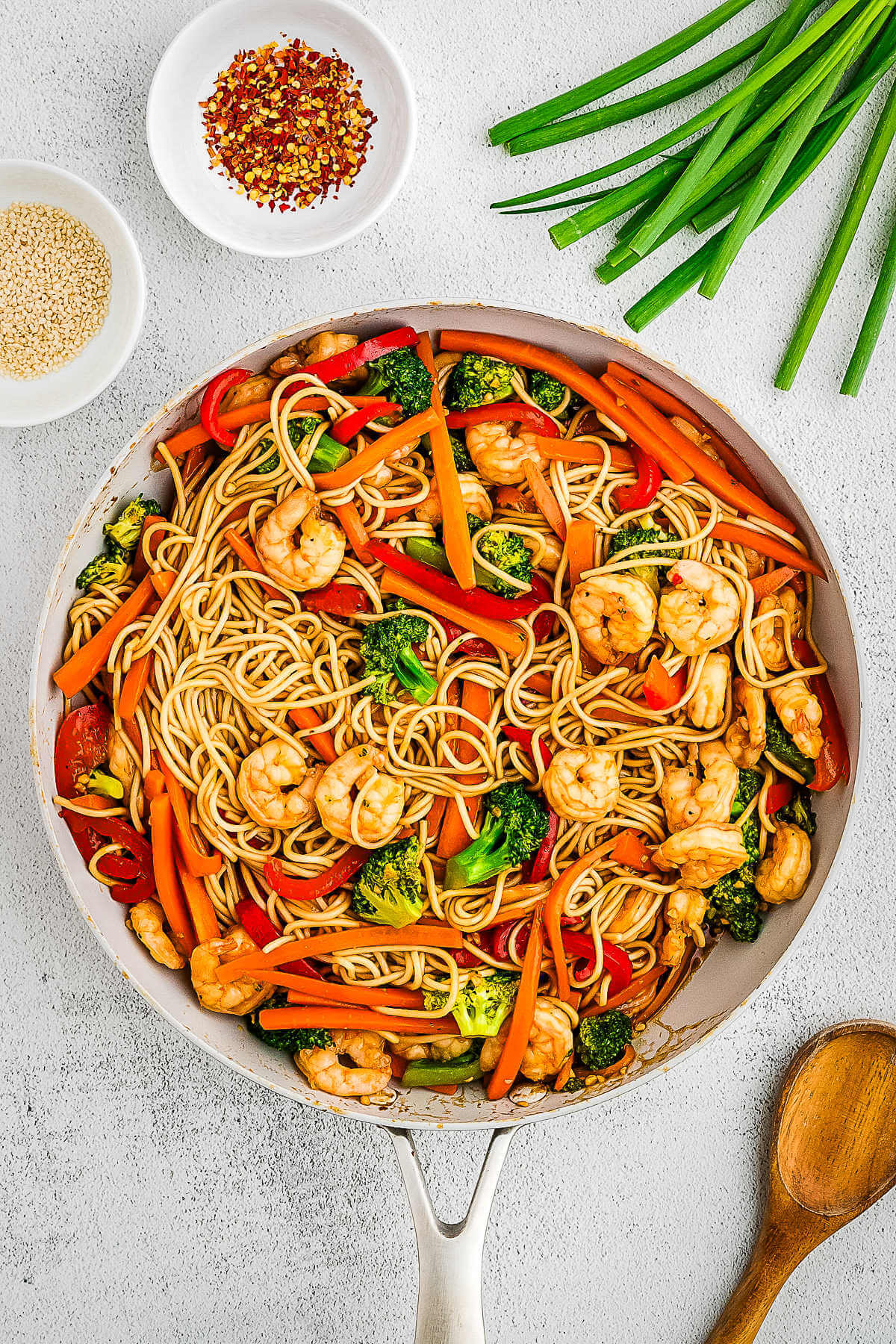 Shrimp Lo Mein in a skillet on a table with green onions and red pepper flakes in the background.