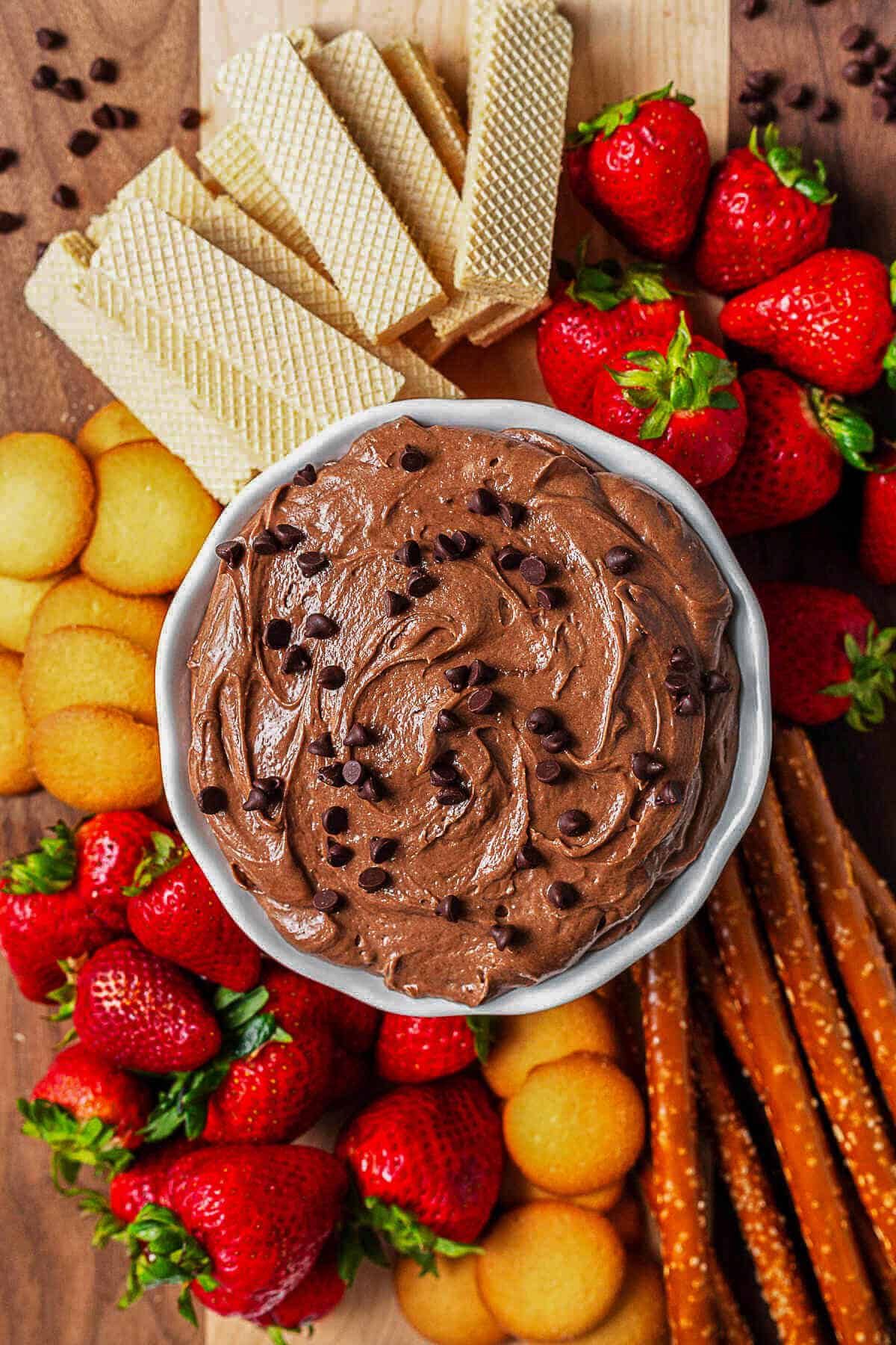 Brownie Batter Dip in a bowl on a wooden board surrounded by strawberries, nilla wafers, cookies, and pretzel rods for dipping.