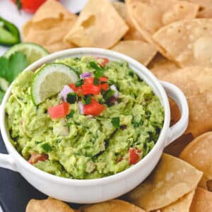 Guacamole garnished with cilantro and chopped tomatoes in a bowl on a table with tortilla chips.