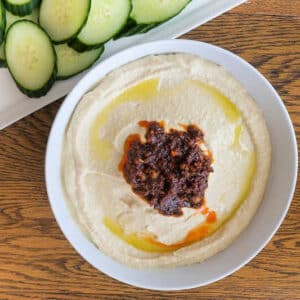 Creamy hummus with spicy walnut topping in a bowl with a plate of cucumbers on a table.