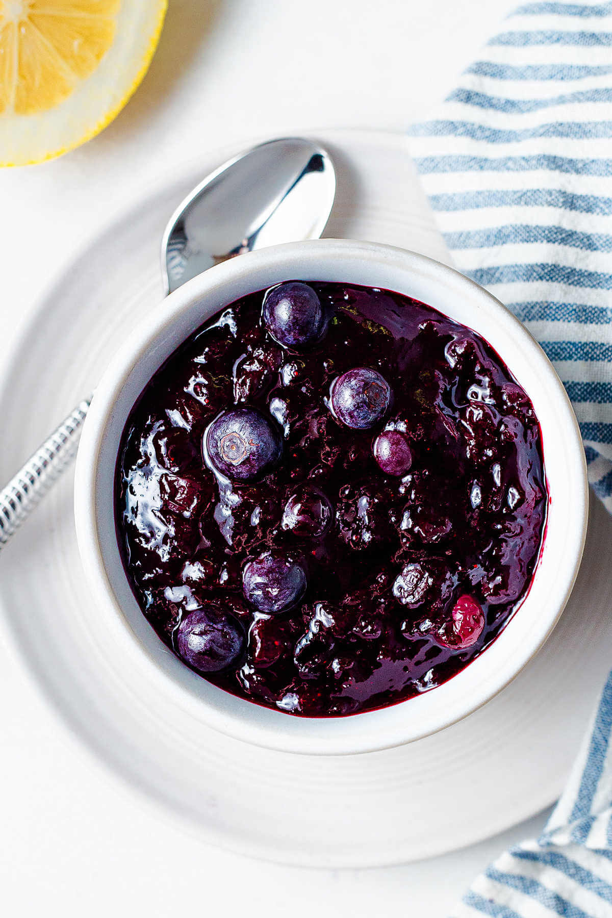 A bowl of blueberry compote on a table with a spoon and lemon half.