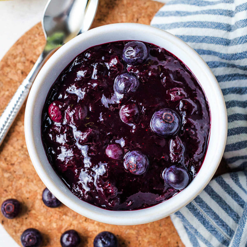 Blueberry compote in a white bowl with blueberries scattered on the table.