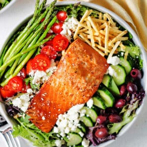 A bowl of salad with grilled salmon on top, asparagus, tomatoes, olives, and feta cheese setting on a table with a cloth napkin.