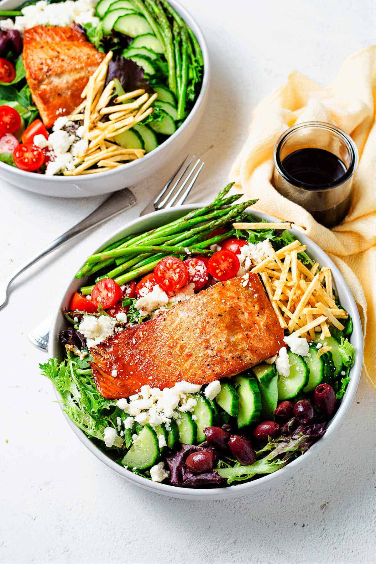 Two plates of salmon and salad on a table with a jar of balsamic dressing, napkins, and forks.