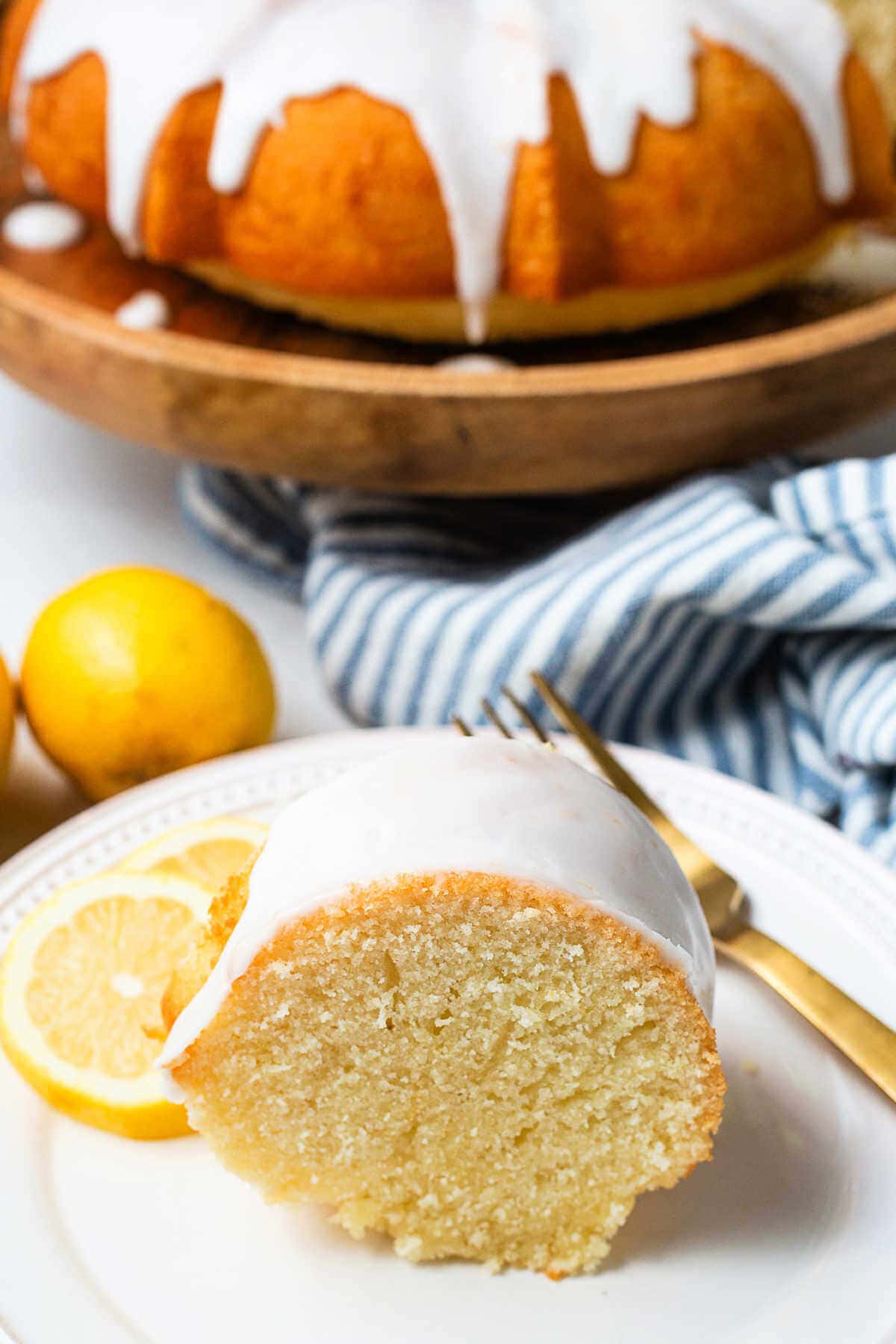 A slice of Lemon Bundt Cake on a plate with lemon slices and a gold fork with the whole cake on a stand in the backtround.