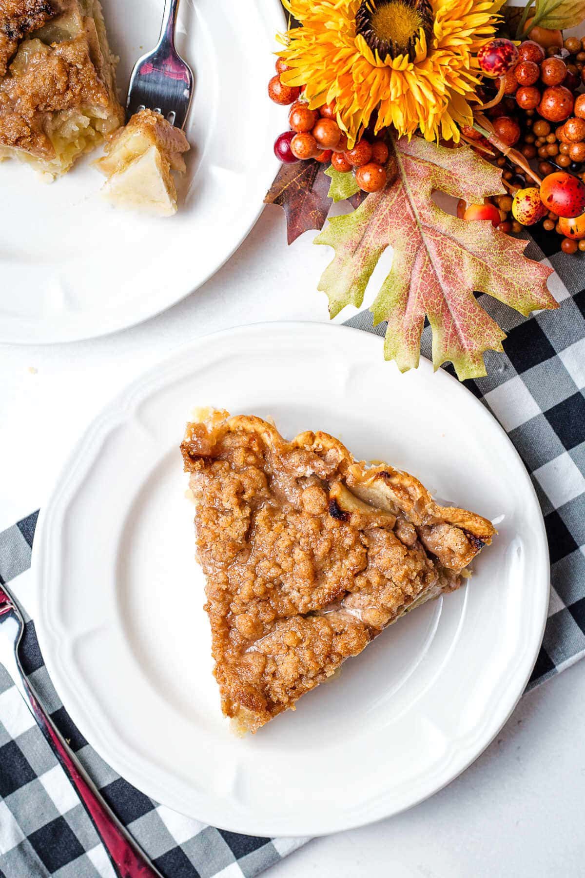 A slice of apple pie with a crumb topping on a plate on a table with fall decor.