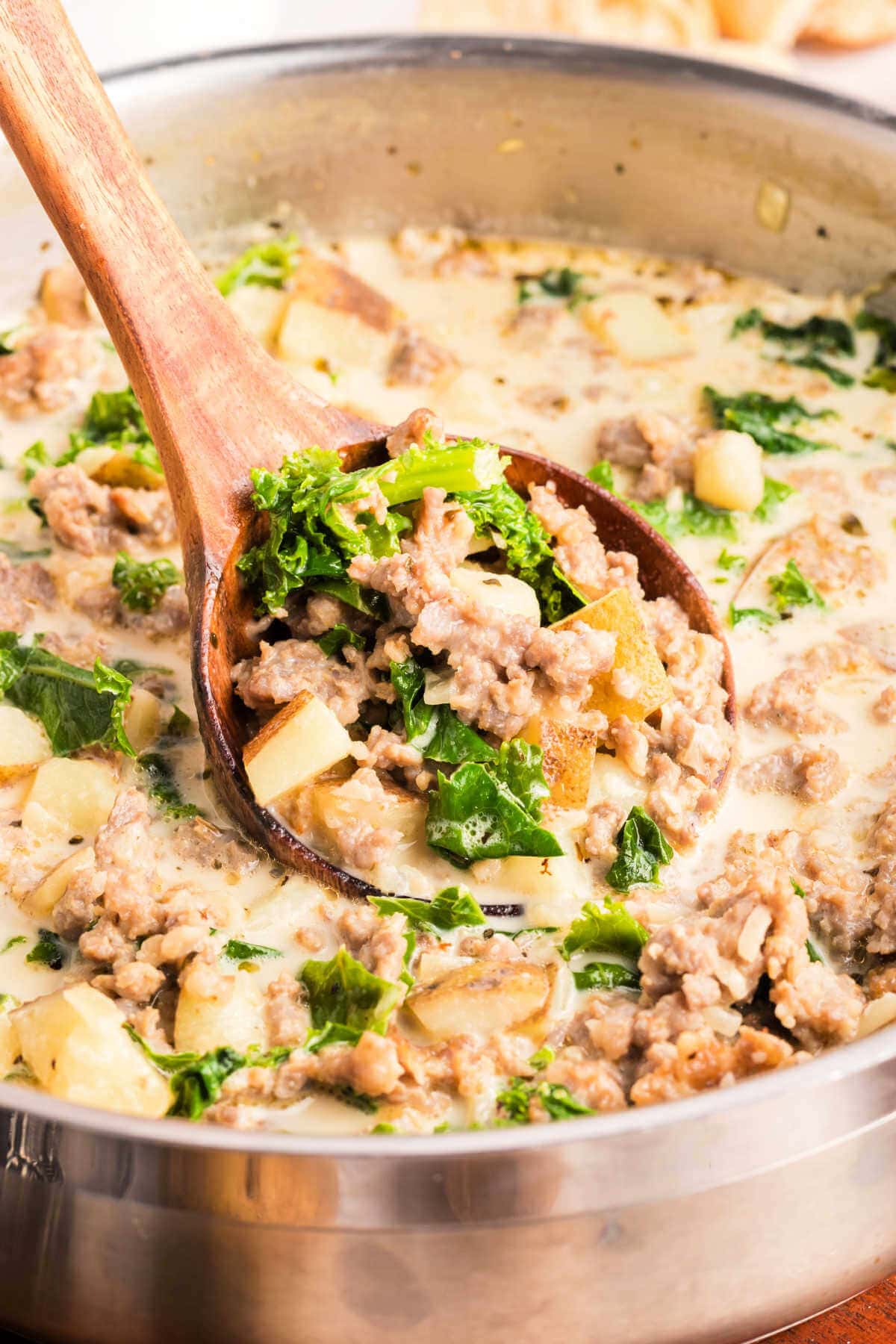 A wooden spoon lifting out a scoop of tuscan soup with kale and potatoes from a large pot.