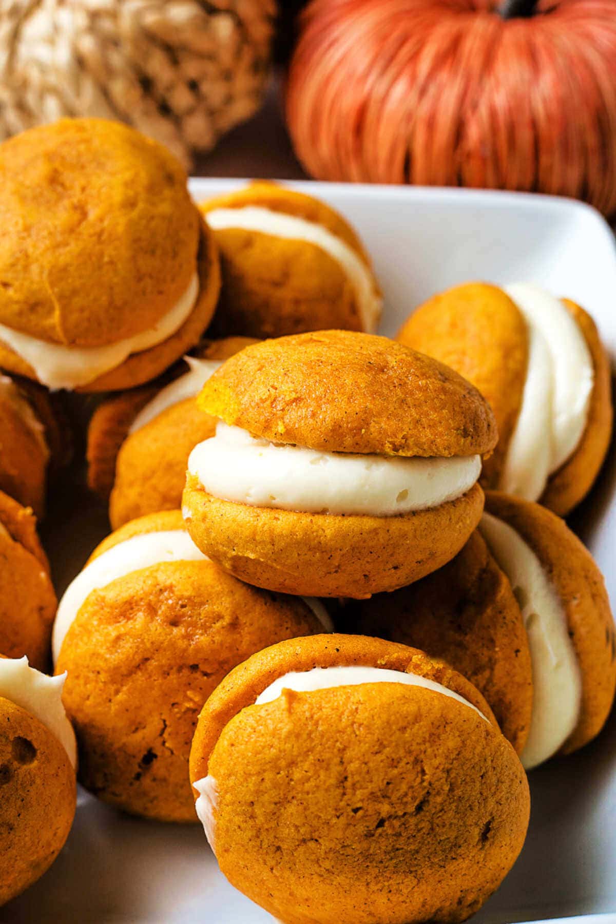 Pumpkin whoopie pies piled into a serving bowl on a table with pumpkin decor in the background.