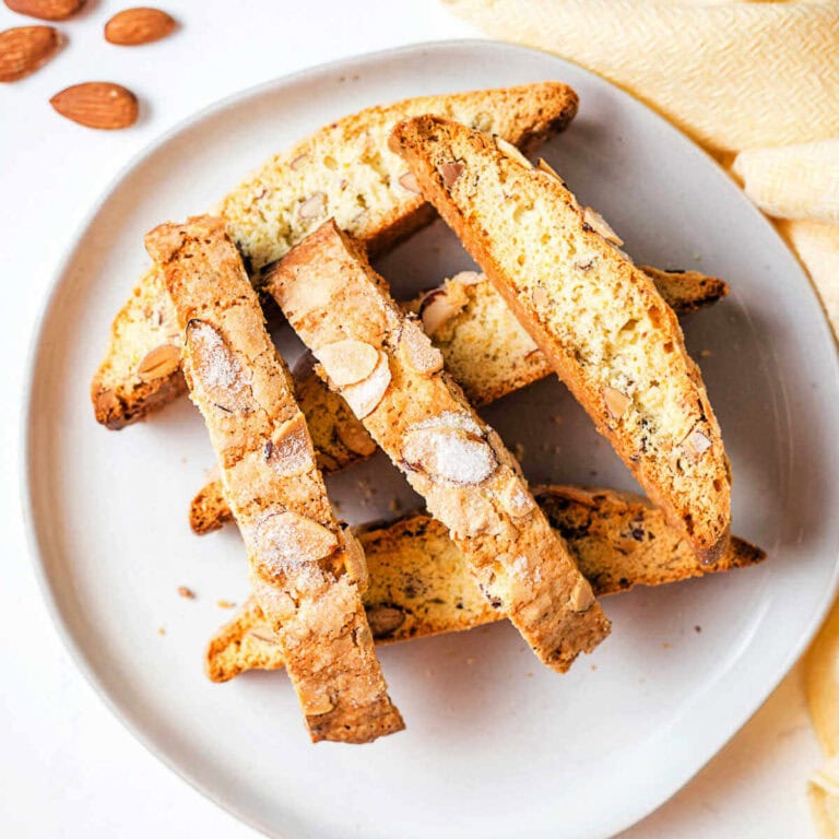 Almond biscotti stacked on a plate with almonds scattered around and a napkin on a table.