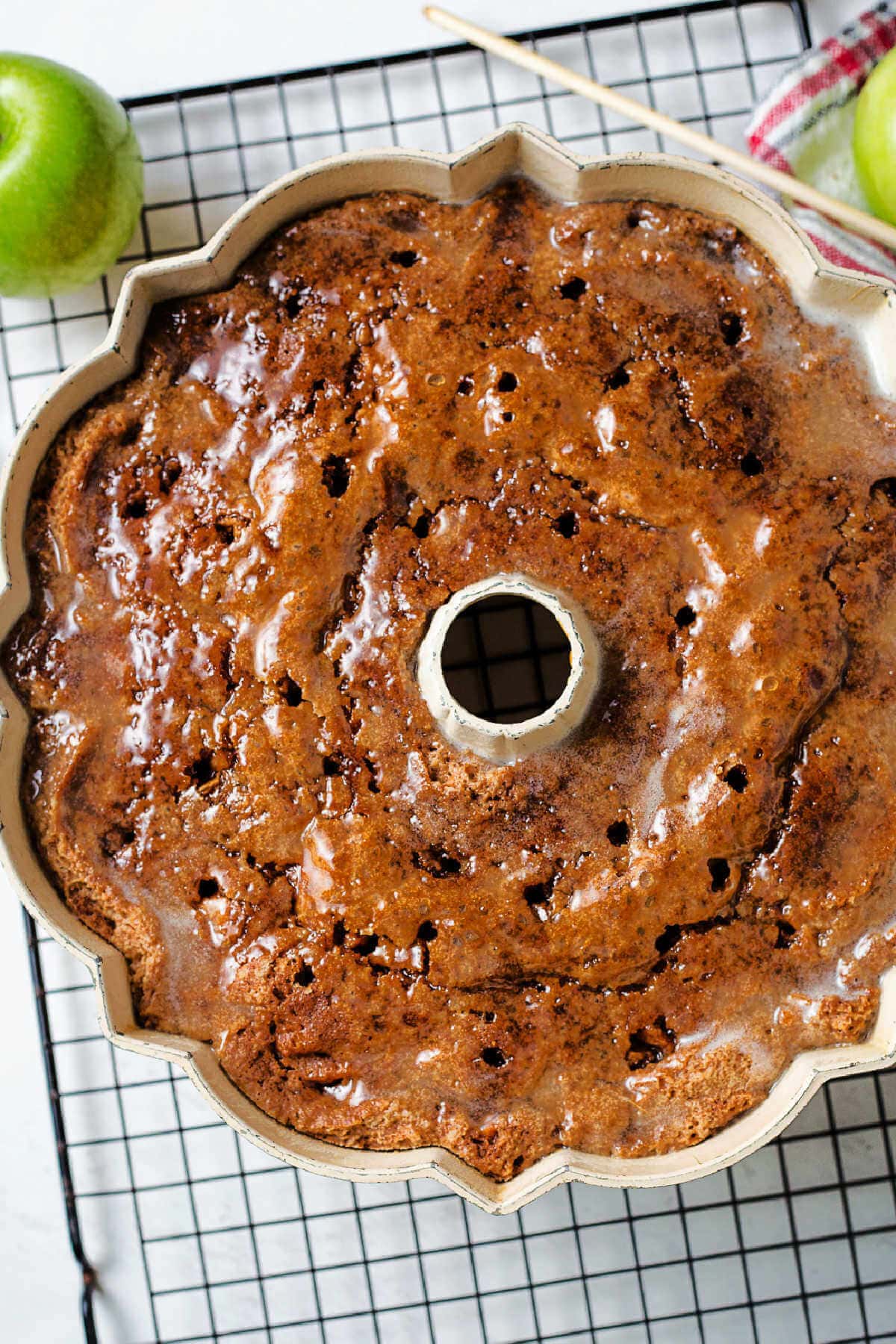 A glazed cake cooling in a pan on a wire rack on a table.