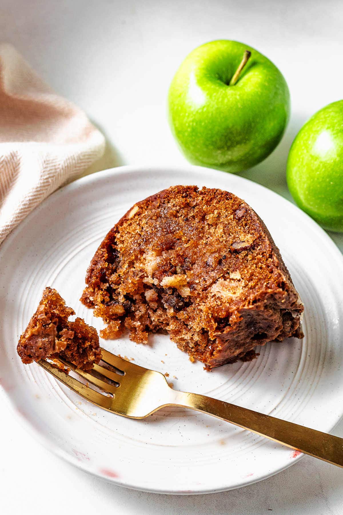 A slice of apple bundt cake on a white plate with a gold fork on a table with granny smith apples in the background.