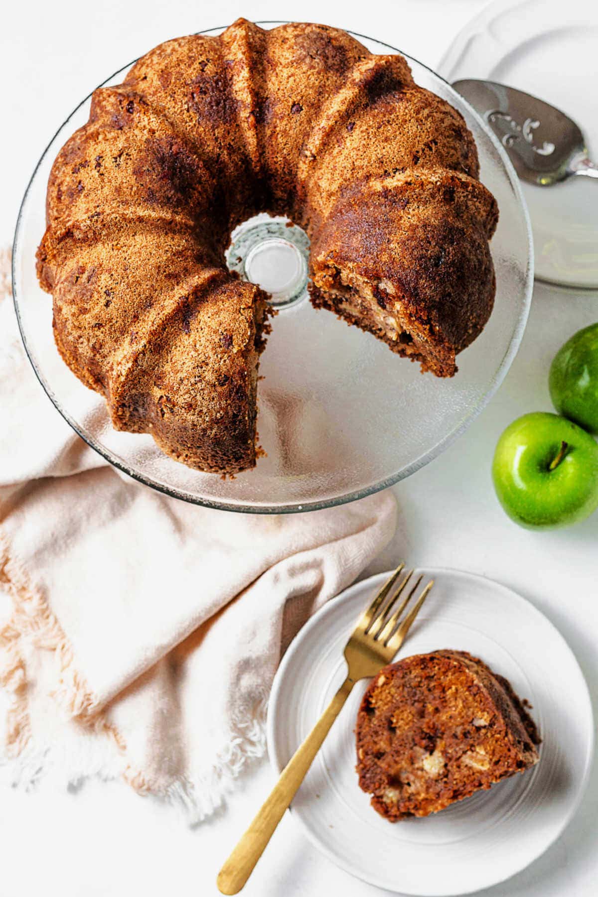 Apple bundt cake on a cake stand on a table with a slice of apple bundt cake on a plate and green apples scattered around.