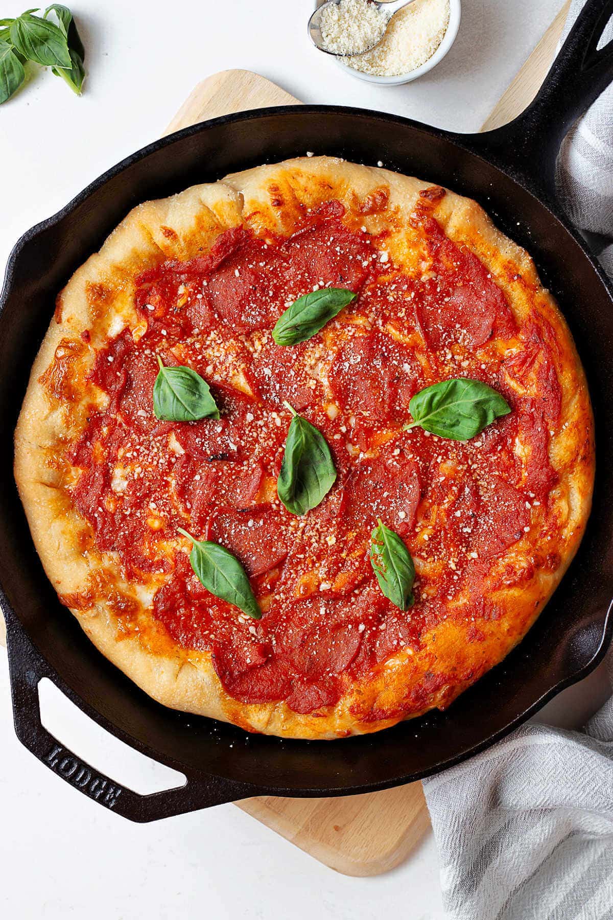 Deep dish pepperoni pizza in a cast iron skillet garnished with fresh basil leaves and grated parmesan cheese on a table.