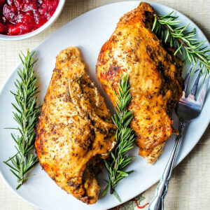 Oven roasted turkey breast removed from the bone and placed on a platter with rosemary sprigs and cranberry sauce on the table.
