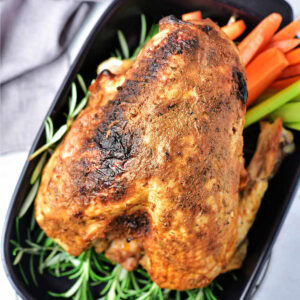 A whole oven roasted turkey breast in a baking dish with fresh herbs and vegetables on a table.