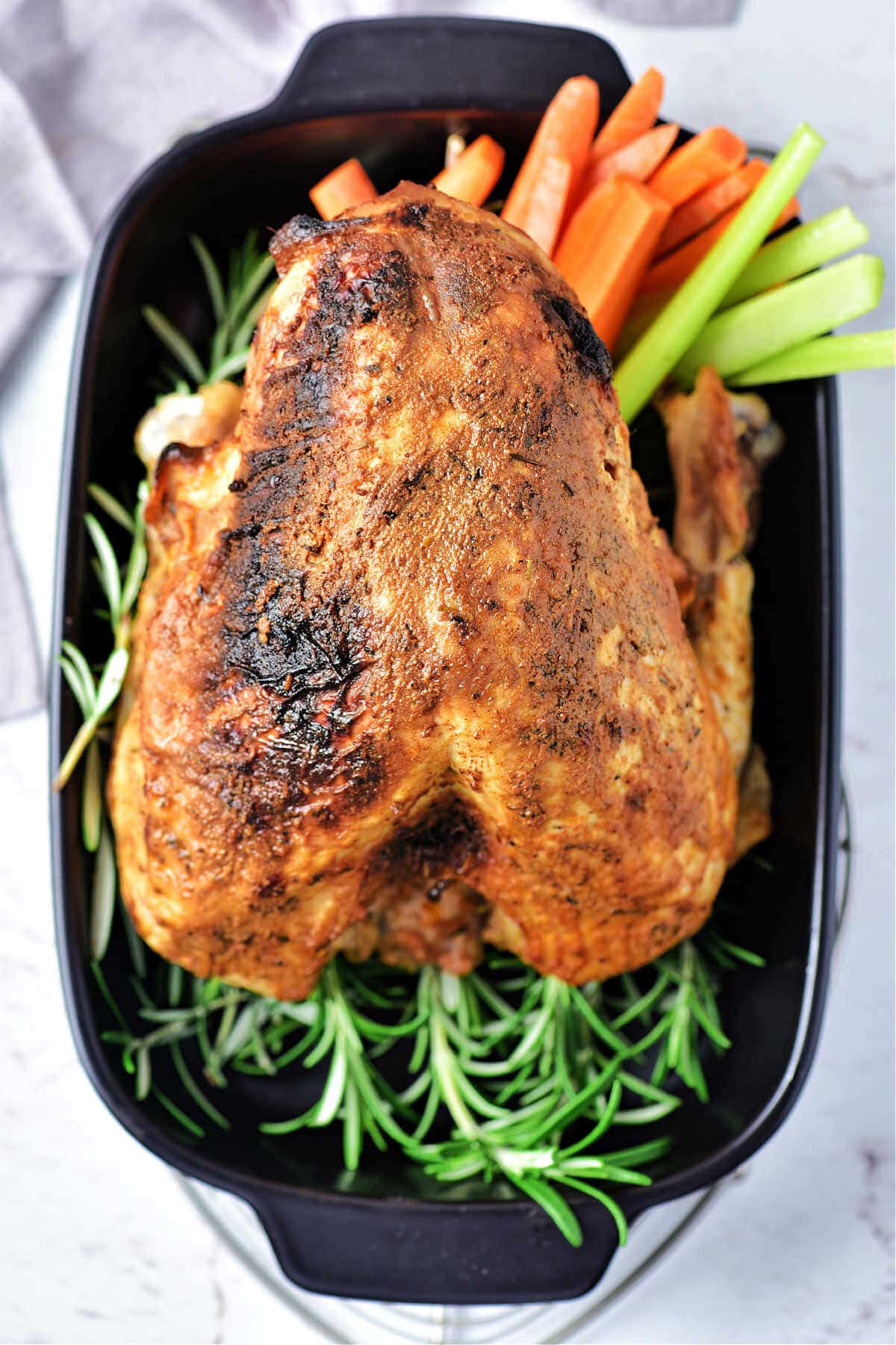 A whole oven roasted turkey breast in a baking dish with fresh herbs and vegetables on a table.