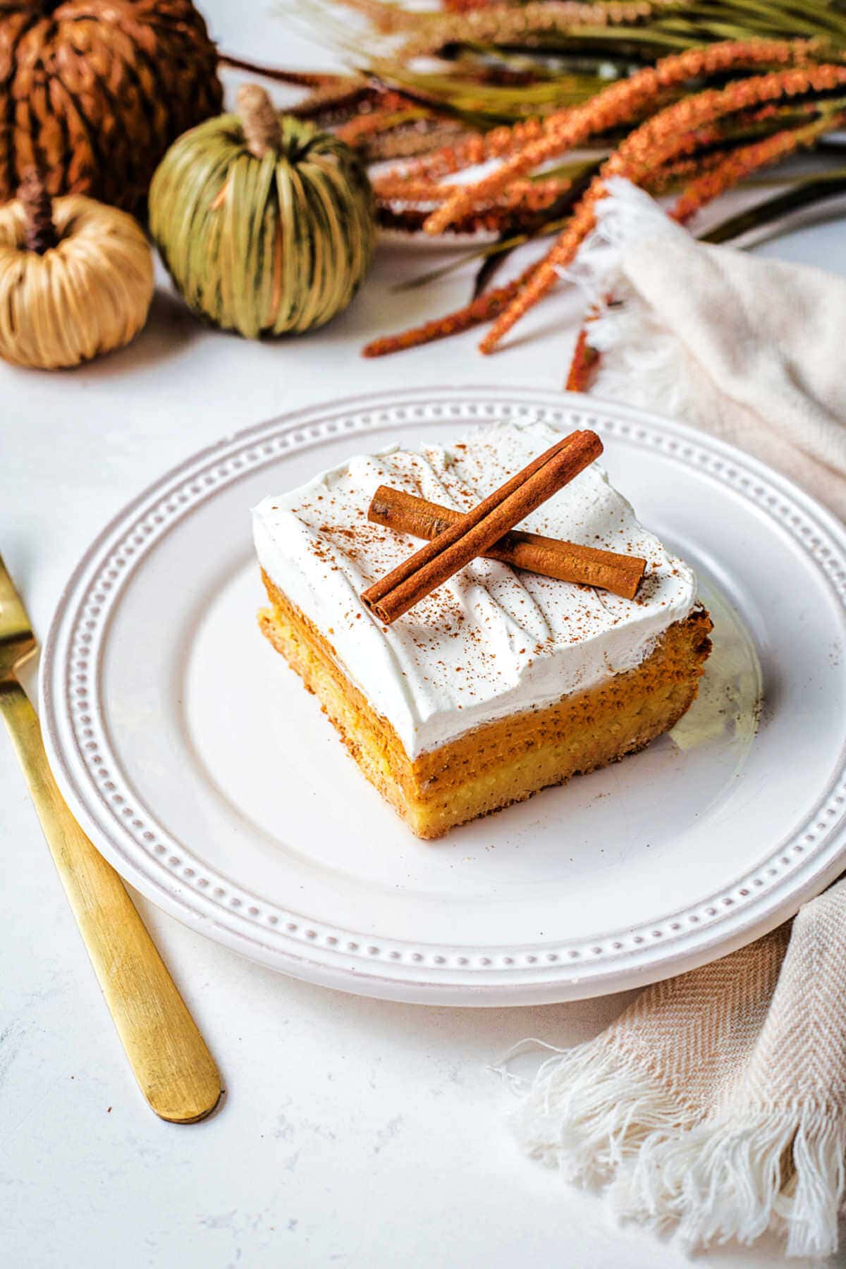 A slice of pumpkin dessert on a plate garnished with cinnamon sticks on a table with Autumn decor in the background.