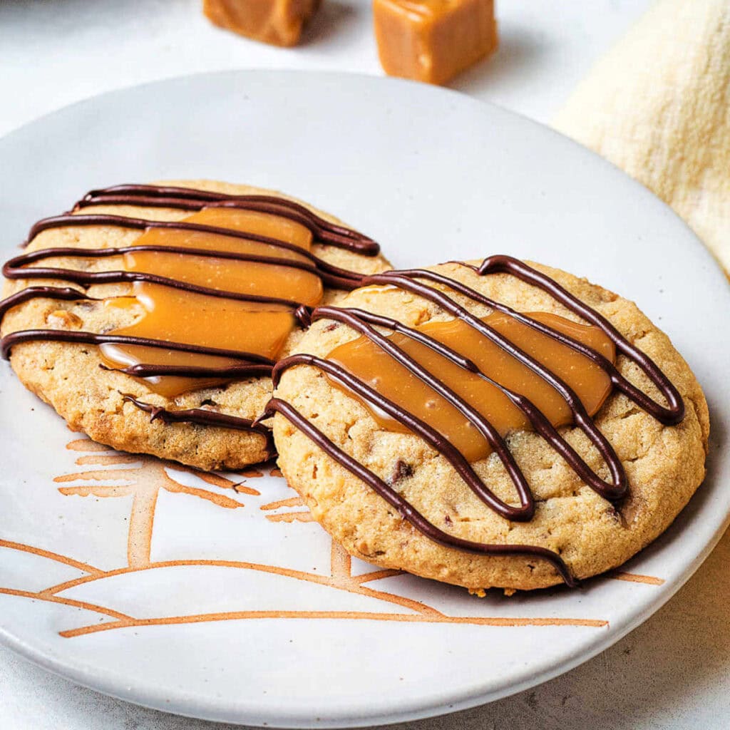 Two Toffee Turtle Cookies with chocolate drizzle on a serving plate.