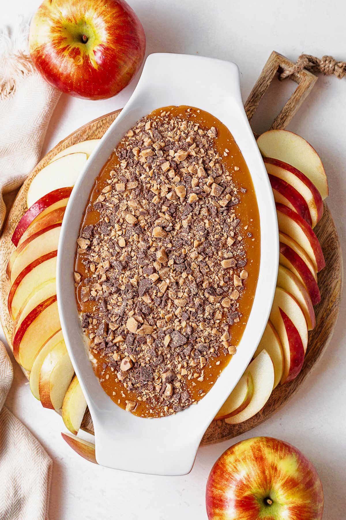 Caramel Apple Dip in a serving dish on a wooden board with apple slices scattered around on a table with extra apples and a linen napkin.