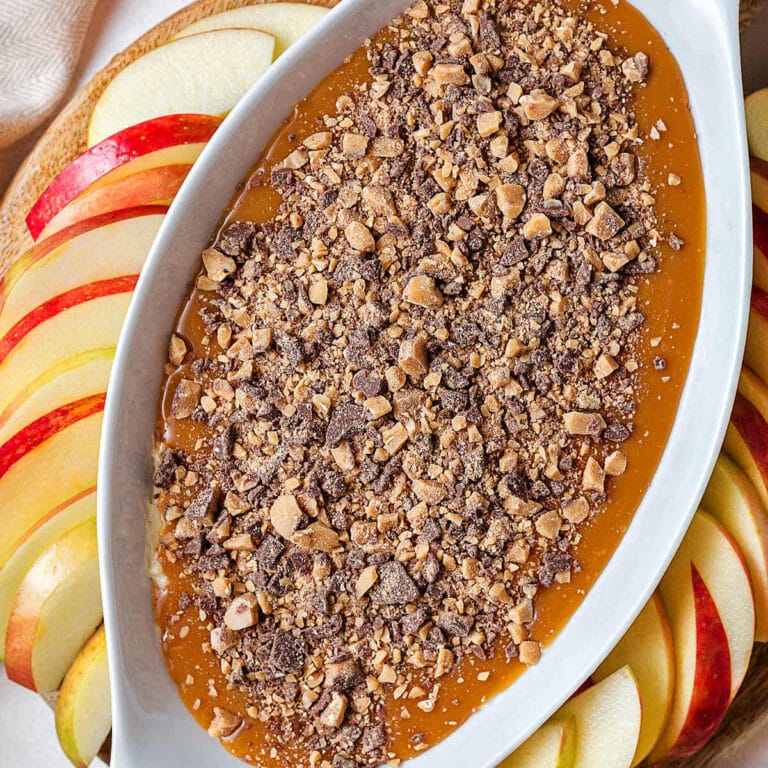 A bowl of Caramel Apple Dip topped with toffee bits on a wooden board with apple slices.