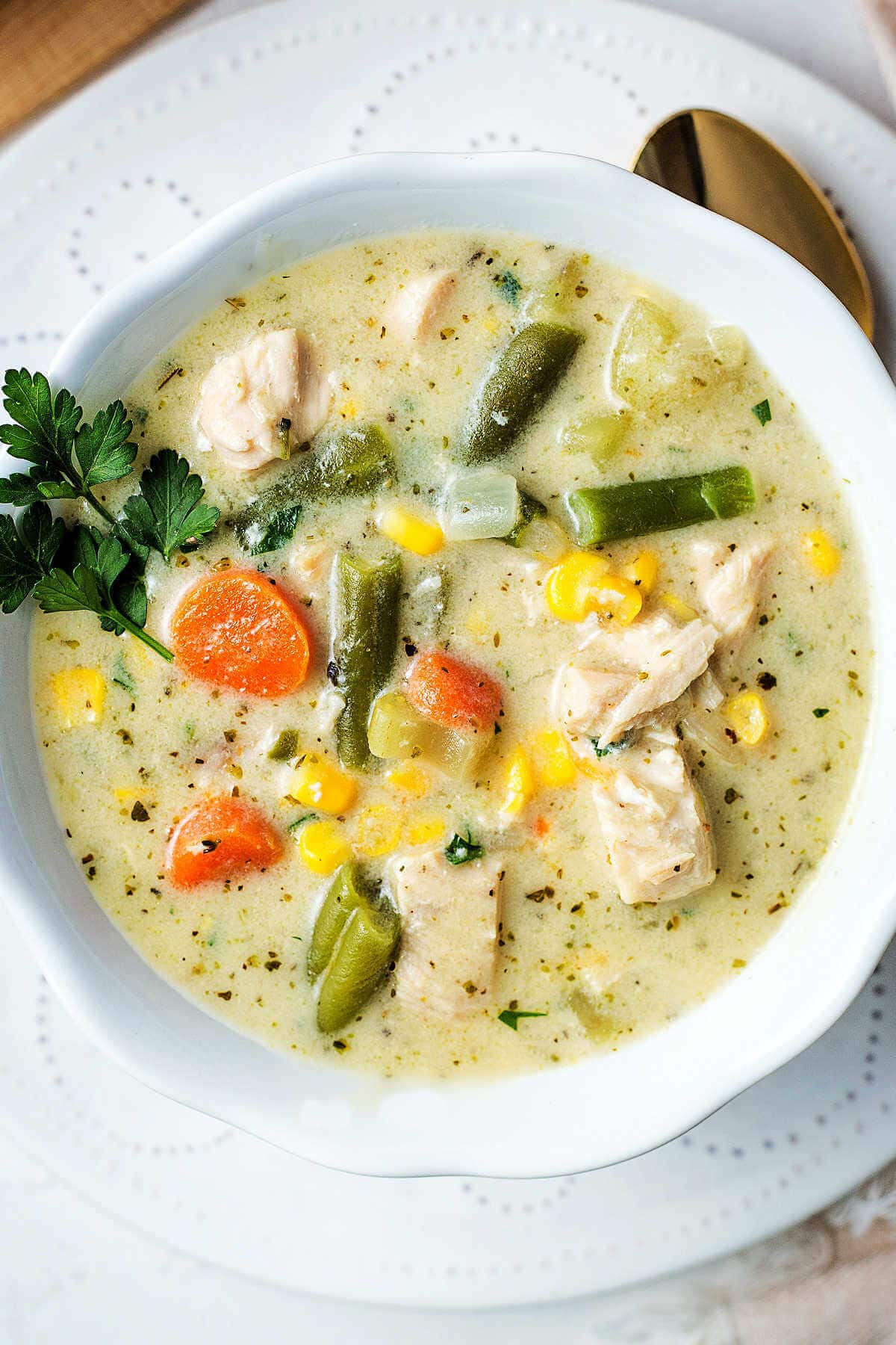 A bowl of soup with chunks of chicken, potatoes, carrots, and green beans on a table garnished with a sprig of fresh parsley.