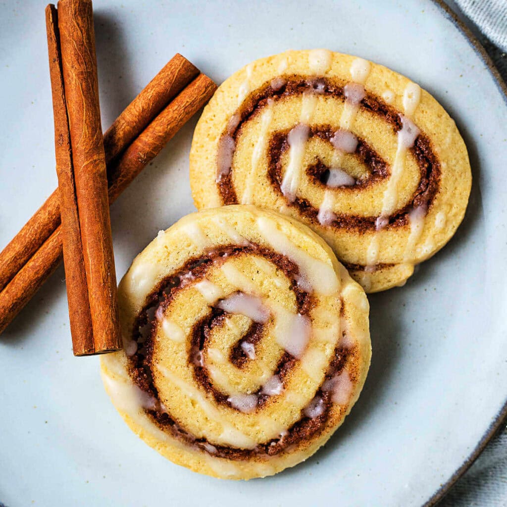 Two cinnamon roll cookies and cinnamon sticks on a plate on a table.