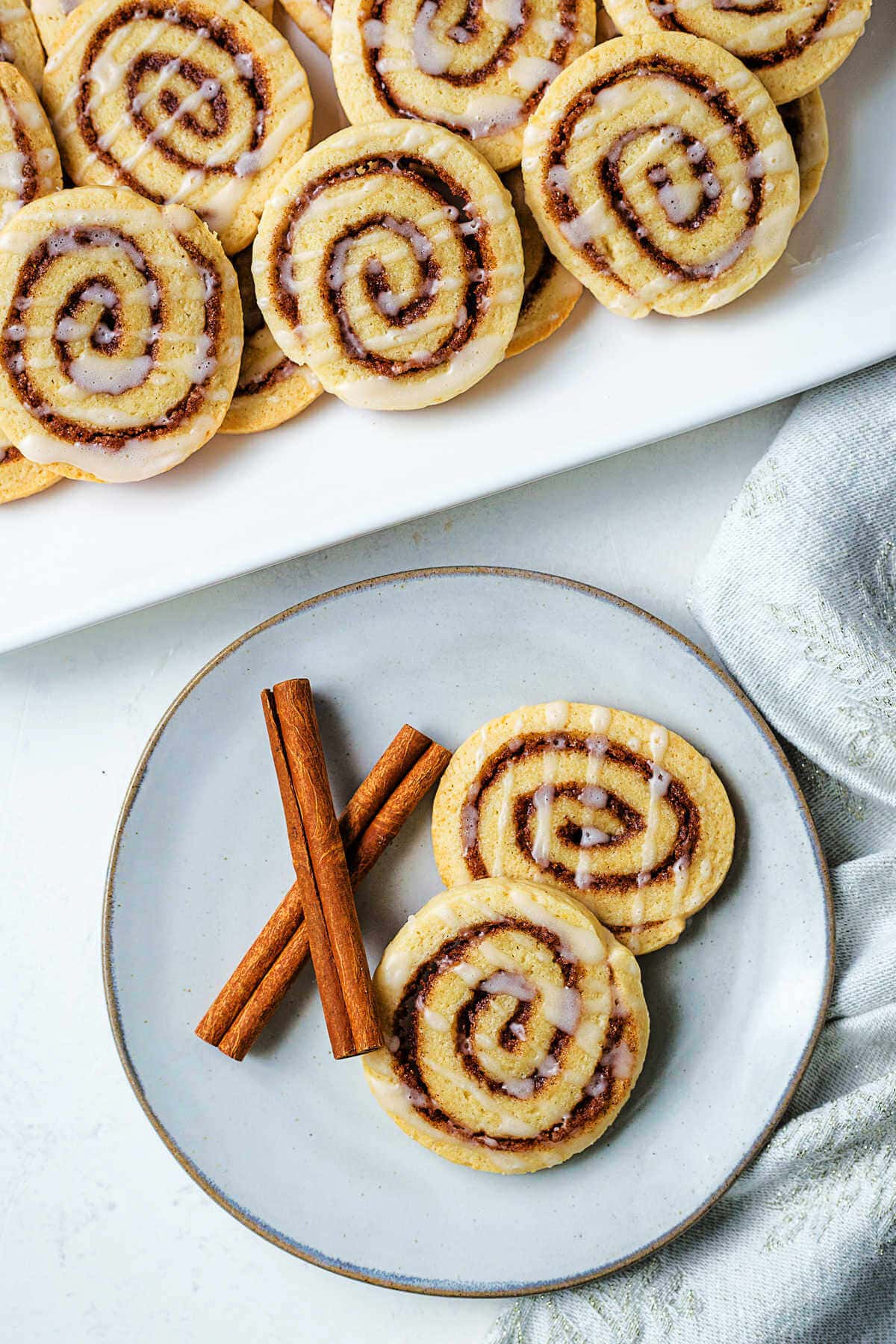 Two cinnamon roll cookies and cinnamon sticks on a plate with a platter of cookies in the background on a table.