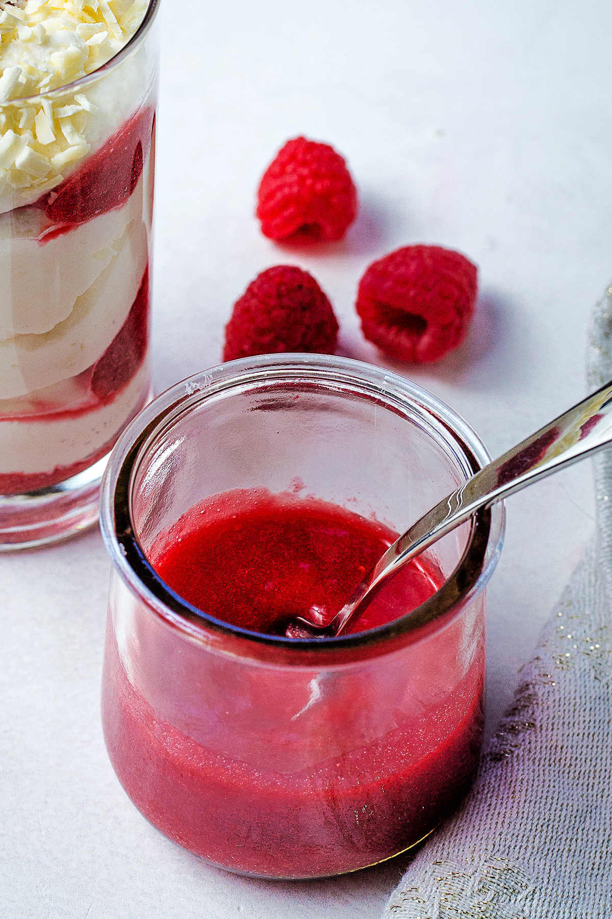 Strained raspberry dessert sauce in a small jar with a spoon on a table with raspberries in the background.