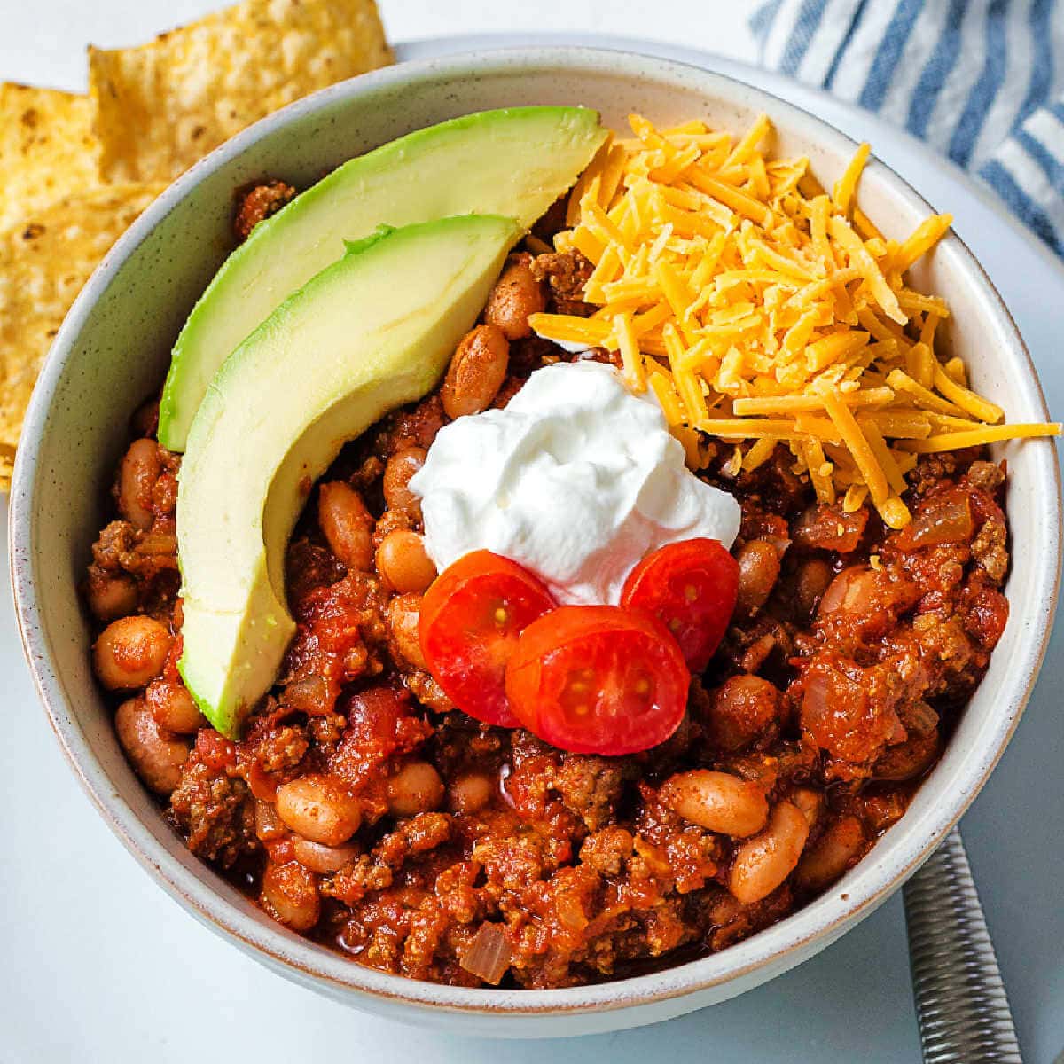 A bowl of red chili garnished with avocado slices, sour cream, diced tomato, and shredded cheese on a table.