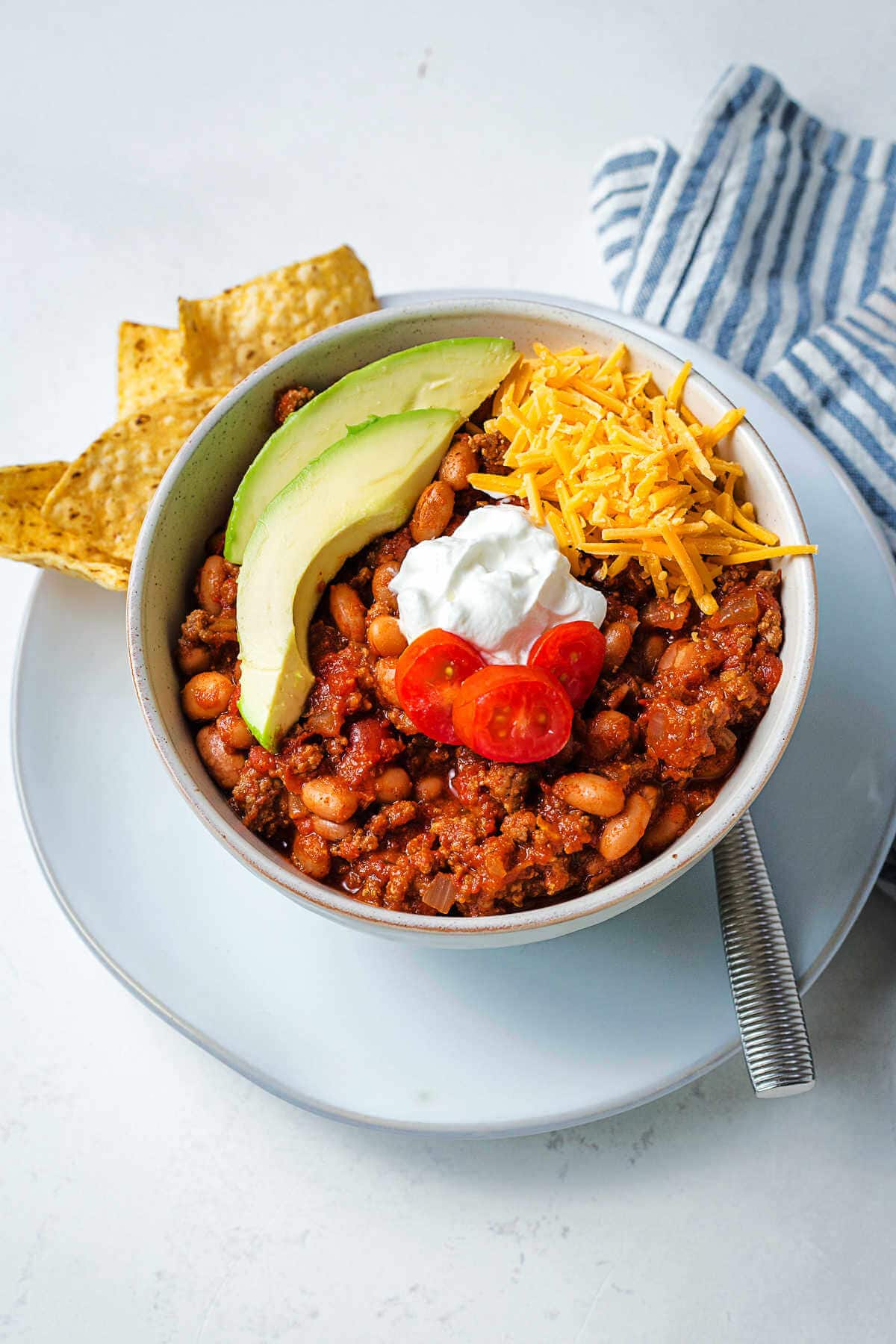 A bowl of red chili garnished with avocado slices, sour cream, diced tomato, and shredded cheese on a plate with tortilla chips sitting on a table.