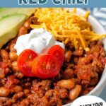 A bowl of red chili garnished with avocado slices, sour cream, diced tomato, and shredded cheese on a table.