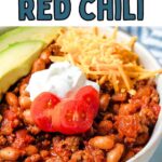 A bowl of red chili garnished with avocado slices, sour cream, diced tomato, and shredded cheese on a table.