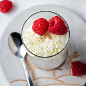 White chocolate mousse in a glass garnished with white chocolate shavings and fresh raspberries on a plate with a spoon on a table.