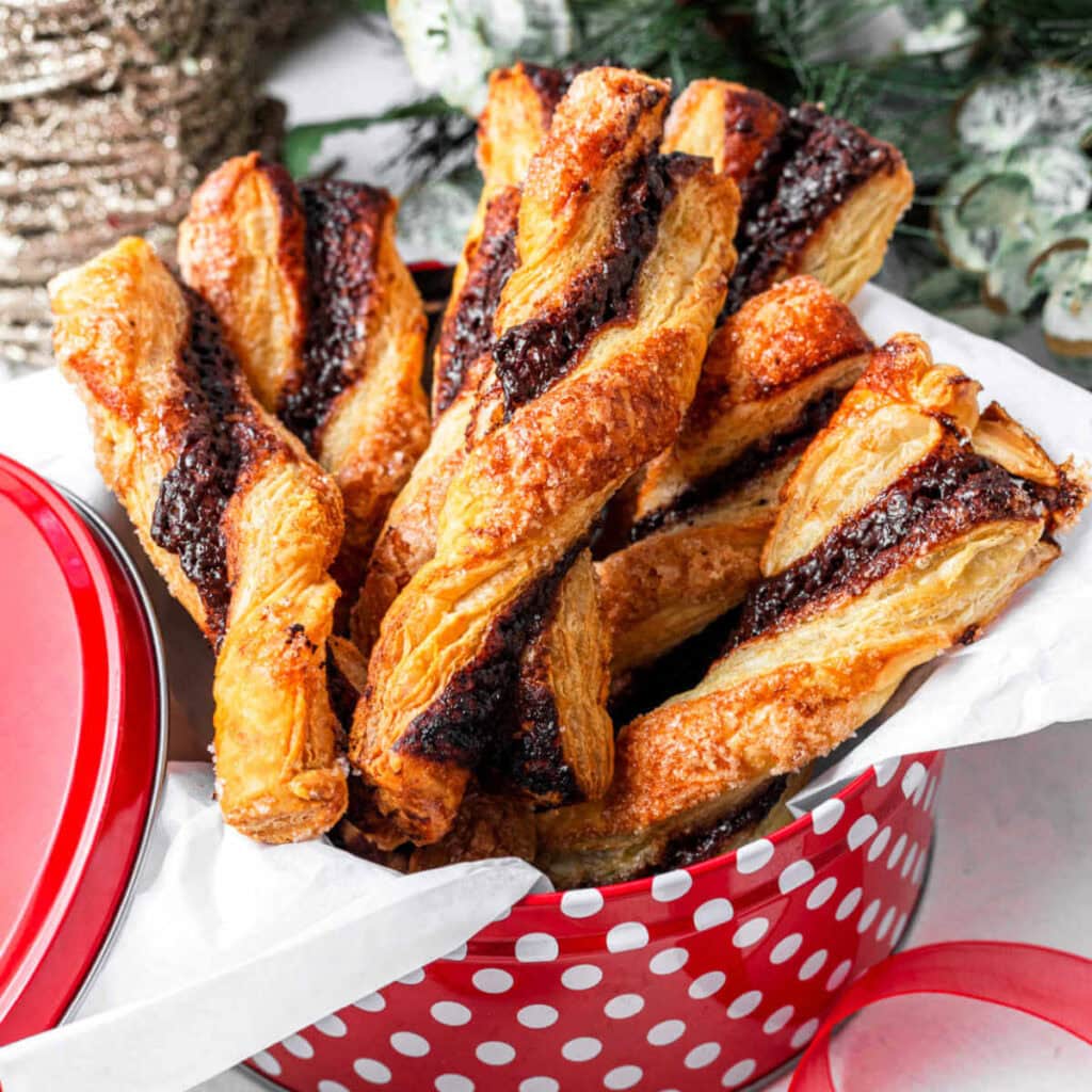 Puff Pastry Chocolate Twists in a holiday tin on a table with Christmas decor in the background.