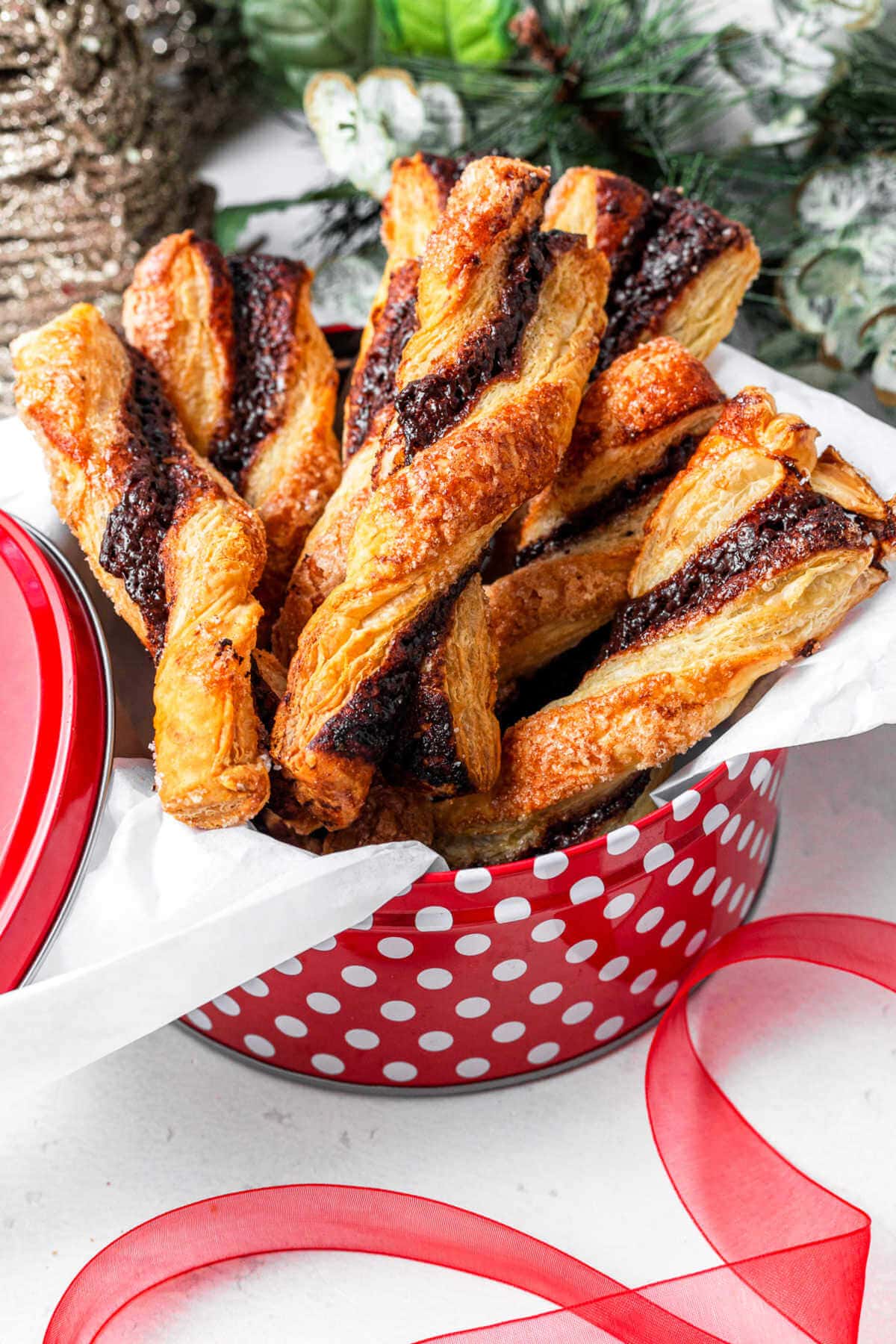 Puff Pastry Chocolate Twists in a holiday tin on a table with Christmas decor in the background.