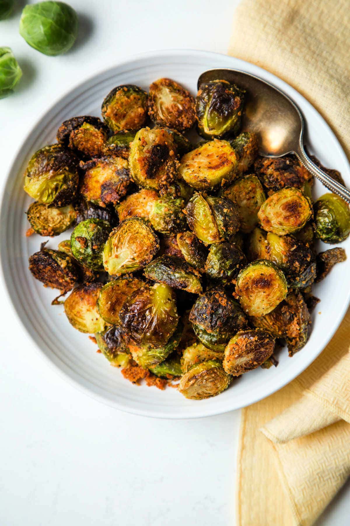 Brussels sprouts in a bowl on a table with a serving spoon and raw Brussels sprouts scattered about.