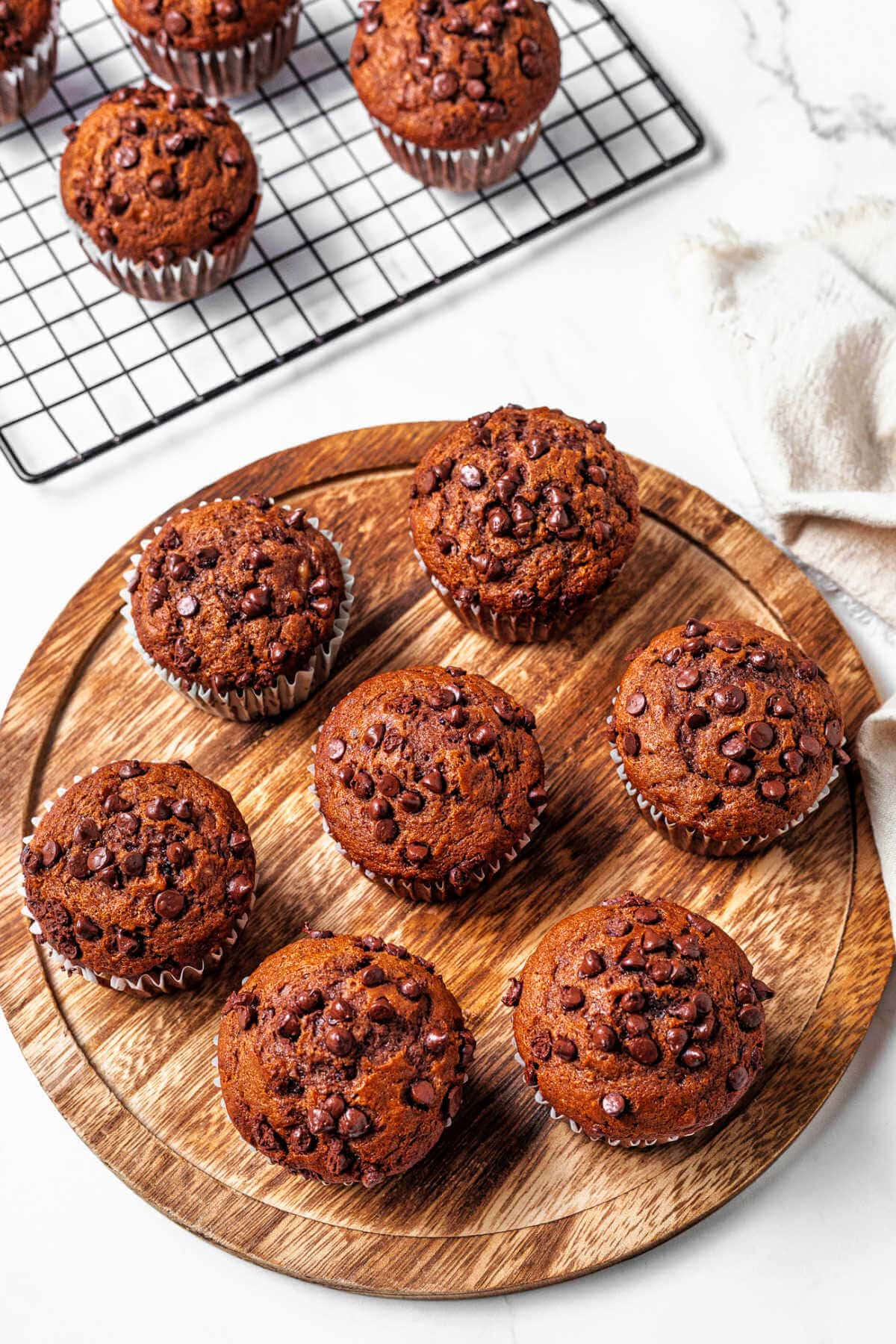 Chocolate Banana Muffins on a wooden board with more muffins in the background on a wire rack on a table.