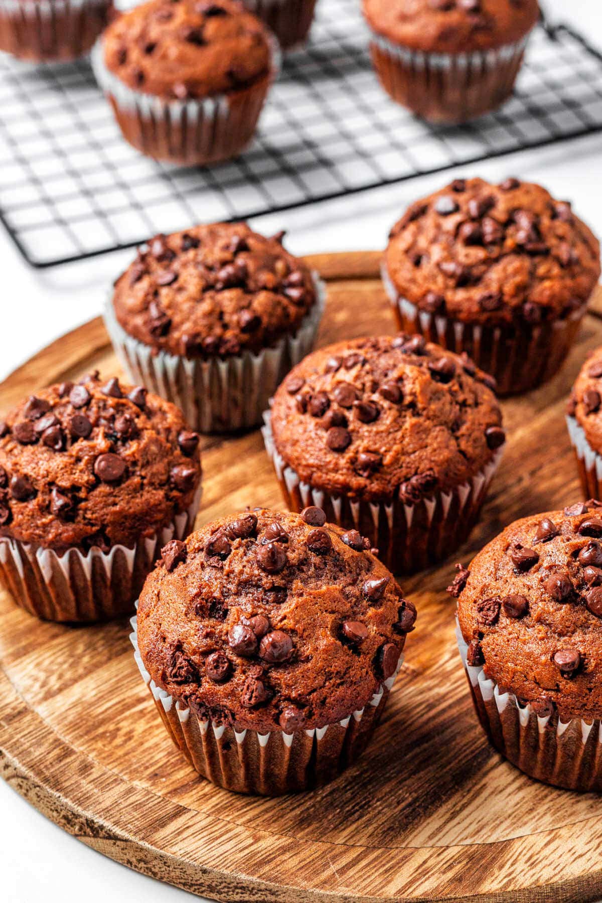 Chocolate Banana Muffins on a wooden board with more muffins in the background on a wire rack on a table.