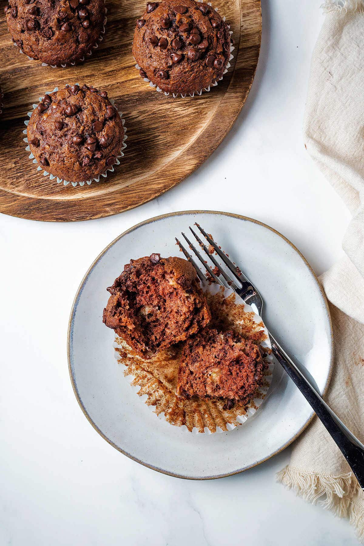 A chocolate banana muffin split in two on a plate with a fork on a table.