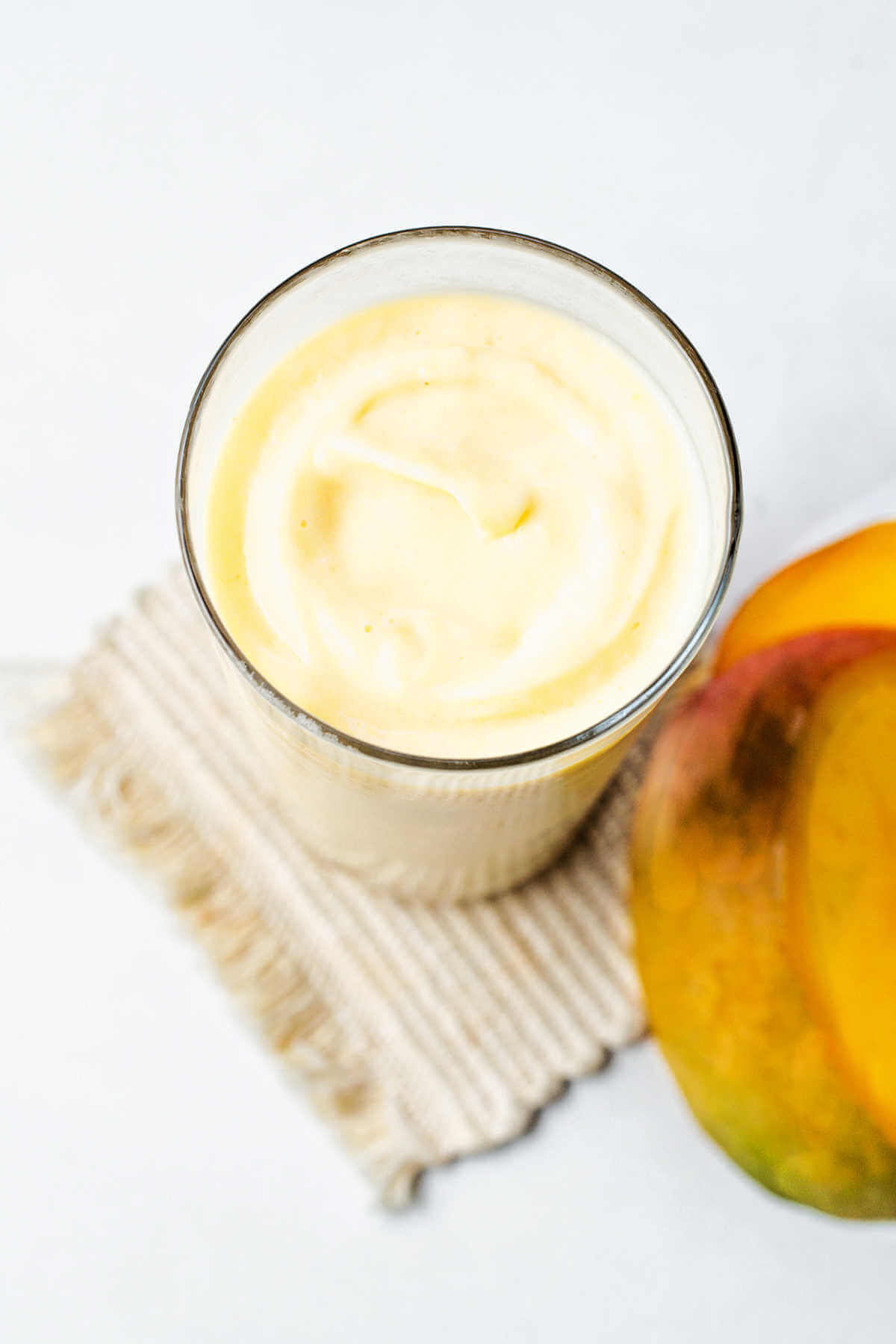 A mango smoothie in a glass on top of a coaster on a table with pieces of mango laying to the side.