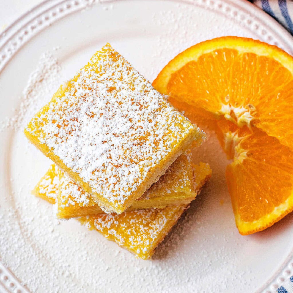 Orange bars stacked on top of each other on a plate with orange slices and powdered sugar dusted on top.