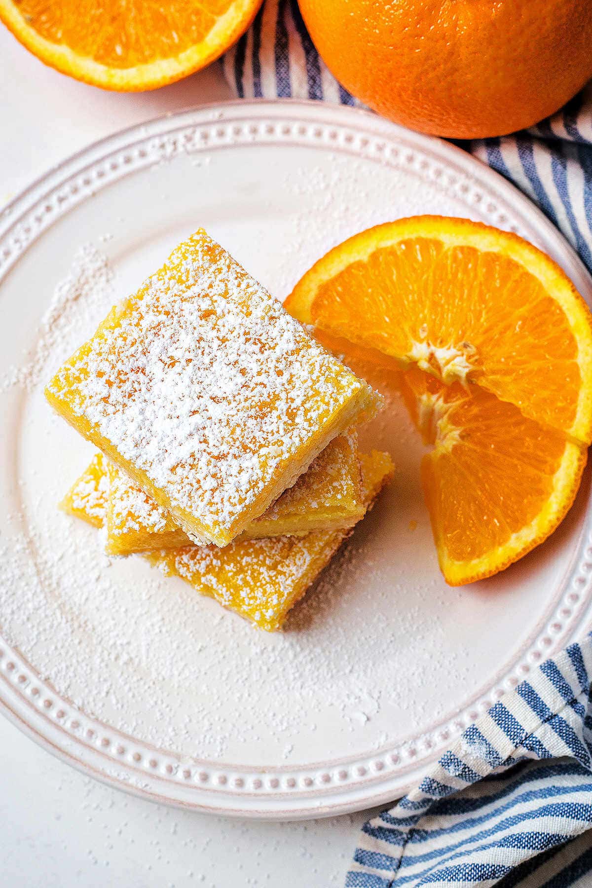 Orange bars stacked on top of each other on a plate with orange slices and powdered sugar dusted on top.