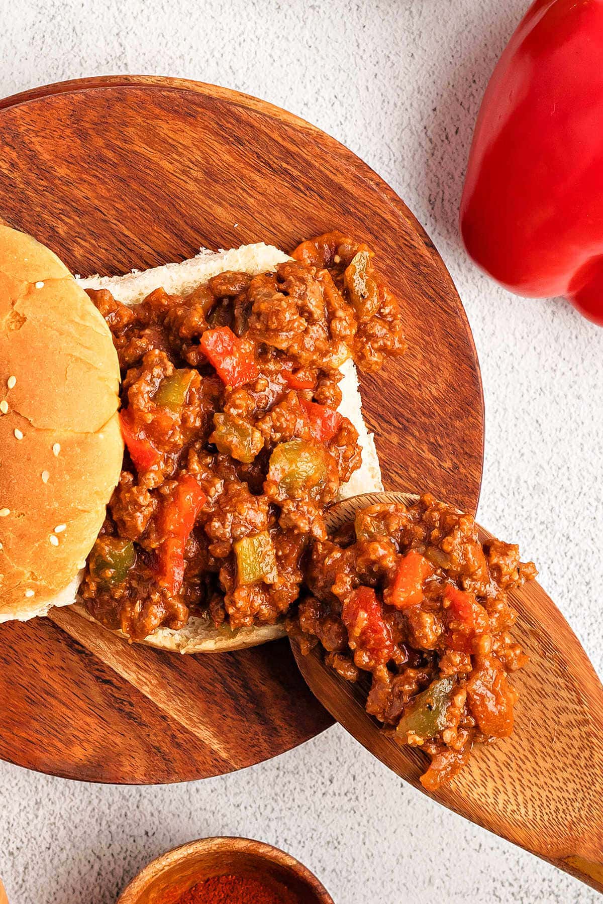 Spooning Sloopy Joe mixture over top a sandwich bun on a wooden board on a table.